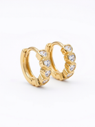 A pair of gold huggie hoop earrings decorated with small clear heart-shaped crystals, shown on a clean white background.